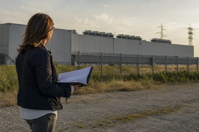Una donna consulta documenti stampati davanti al perimetro recintato di un grande data center al tramonto.