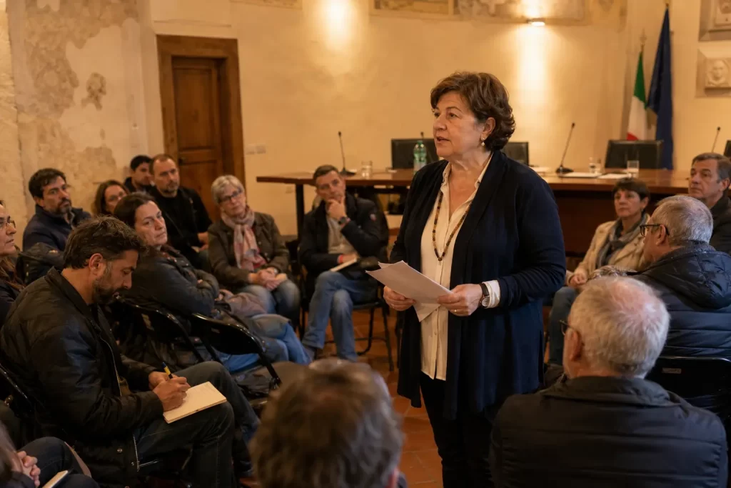 data-center-osservazioni-via-assemblea-pubblica-cittadini - Eywa Divulgazione Una donna interviene in piedi durante un'assemblea pubblica in una sala comunale, con altri cittadini seduti che ascoltano