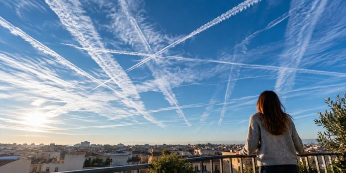 Persona di spalle su un terrazzo guarda un cielo limpido attraversato da scie di aerei incrociate, sopra i tetti di una città